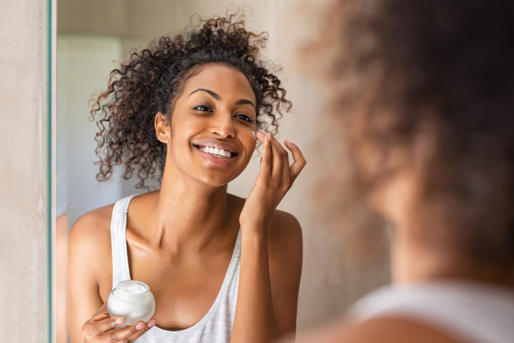 Black girl take care of her beautiful skin. Young african woman applying moisturizer on her face. Smiling black natural girl holding little jar of skin lotion in bathroom for beauty treatment routine.